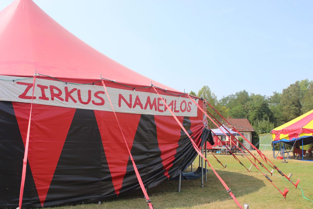 Schwarz-rotes Zirkuszelt mit "Zirkus Namenlos" Banner, im Hintergrund ein weiteres kleines Zirkuszelt und ein Bauwagen zu sehen